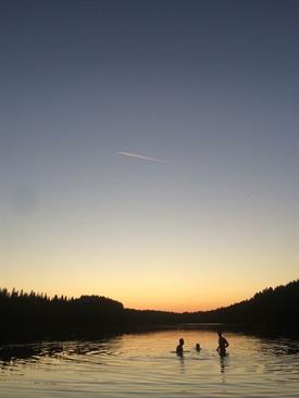 Die Silouhetten von 3 Personen, die Hüft-tief in einem See stehen. Vor Ihnen liegt ein dunkler Wald und ein bunter Sonnenuntergang.